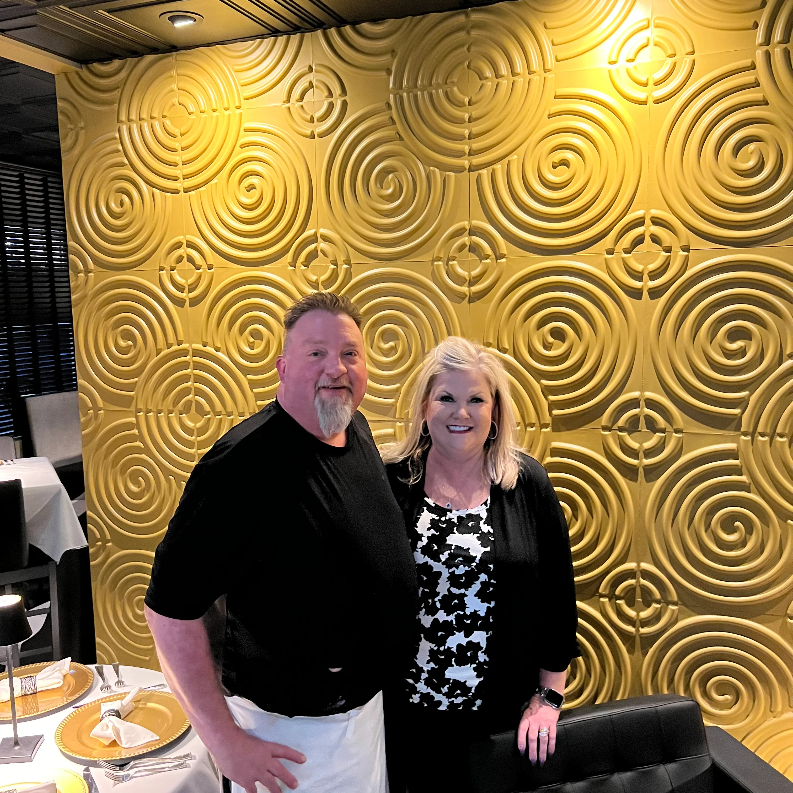 Chef Todd and Nickki Golden stand smiling together in a stylish restaurant setting. Behind them is a striking gold wall with a bold spiral pattern design. The table in front is elegantly set with gold chargers, white napkins, and modern black chairs, creating a warm and upscale dining atmosphere.