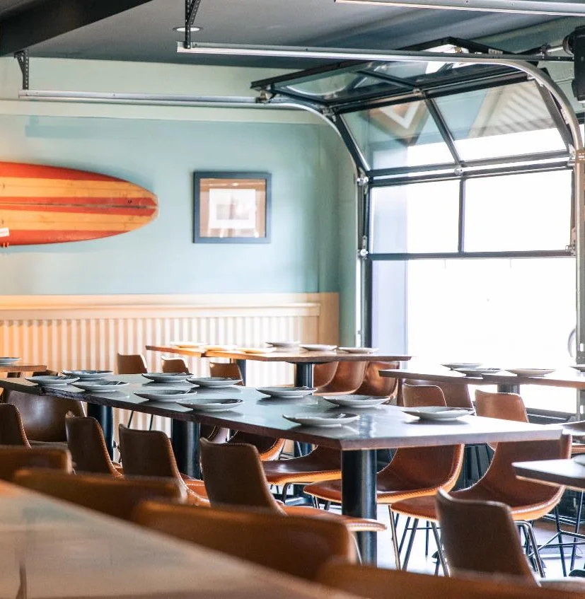 A spacious restaurant interior featuring long wooden tables and leather chairs, with a surfboard displayed on the wall and large garage-style windows letting in natural light.