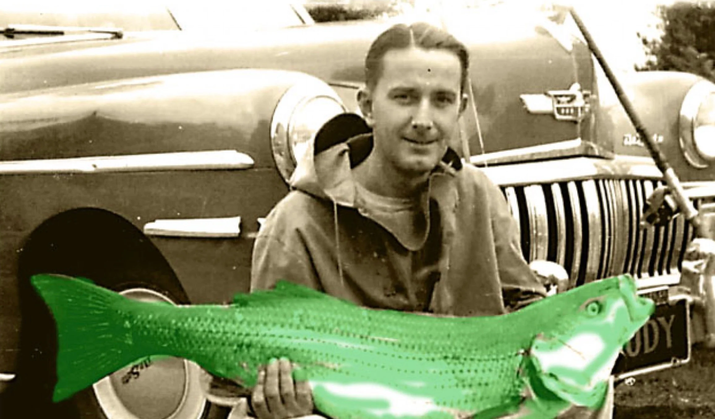 Man holding a large green fish near a vintage car
