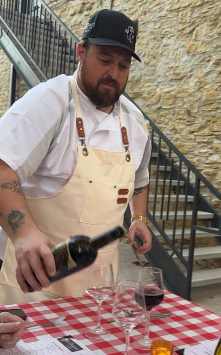 Chef pouring red wine at a rustic outdoor dining table