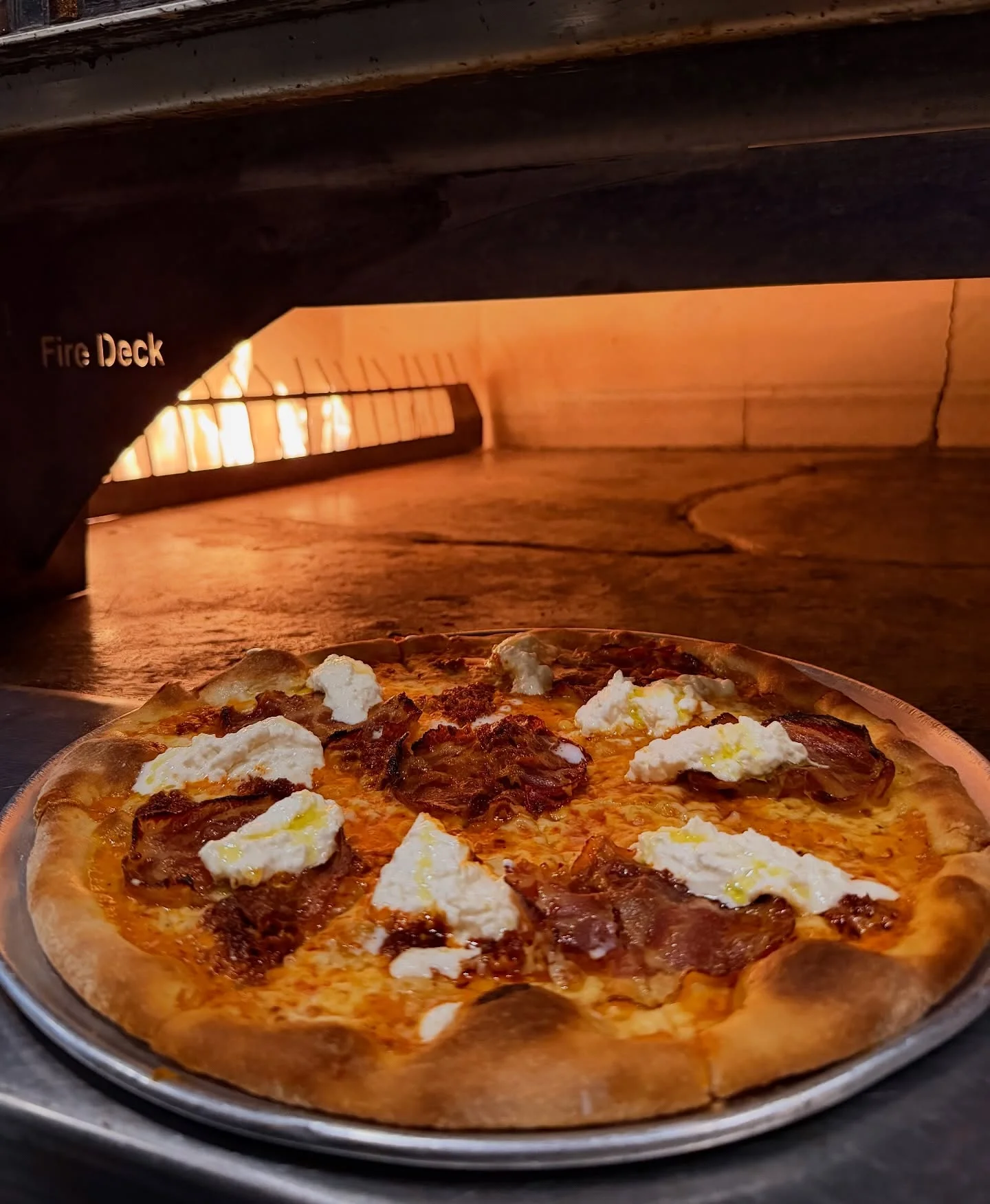 A delicious pizza topped with ricotta and crispy meat, placed on a silver tray in an oven with flames visible in the background.