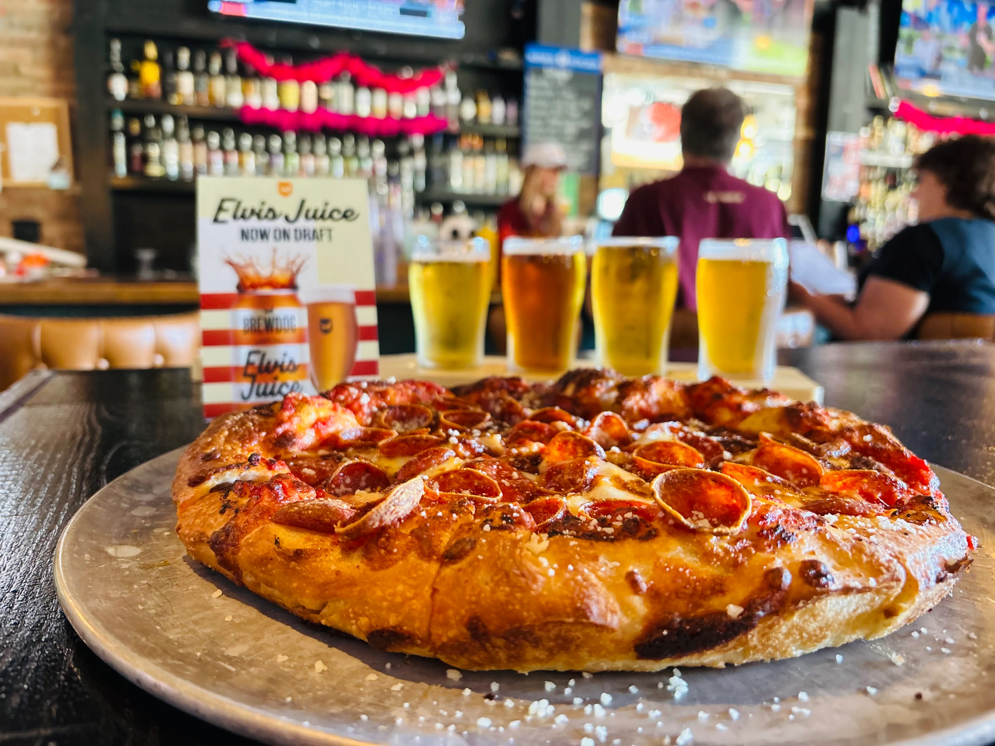 A freshly baked pizza topped with pepperoni, served on a metal tray, accompanied by several glasses of beer in a lively bar setting.