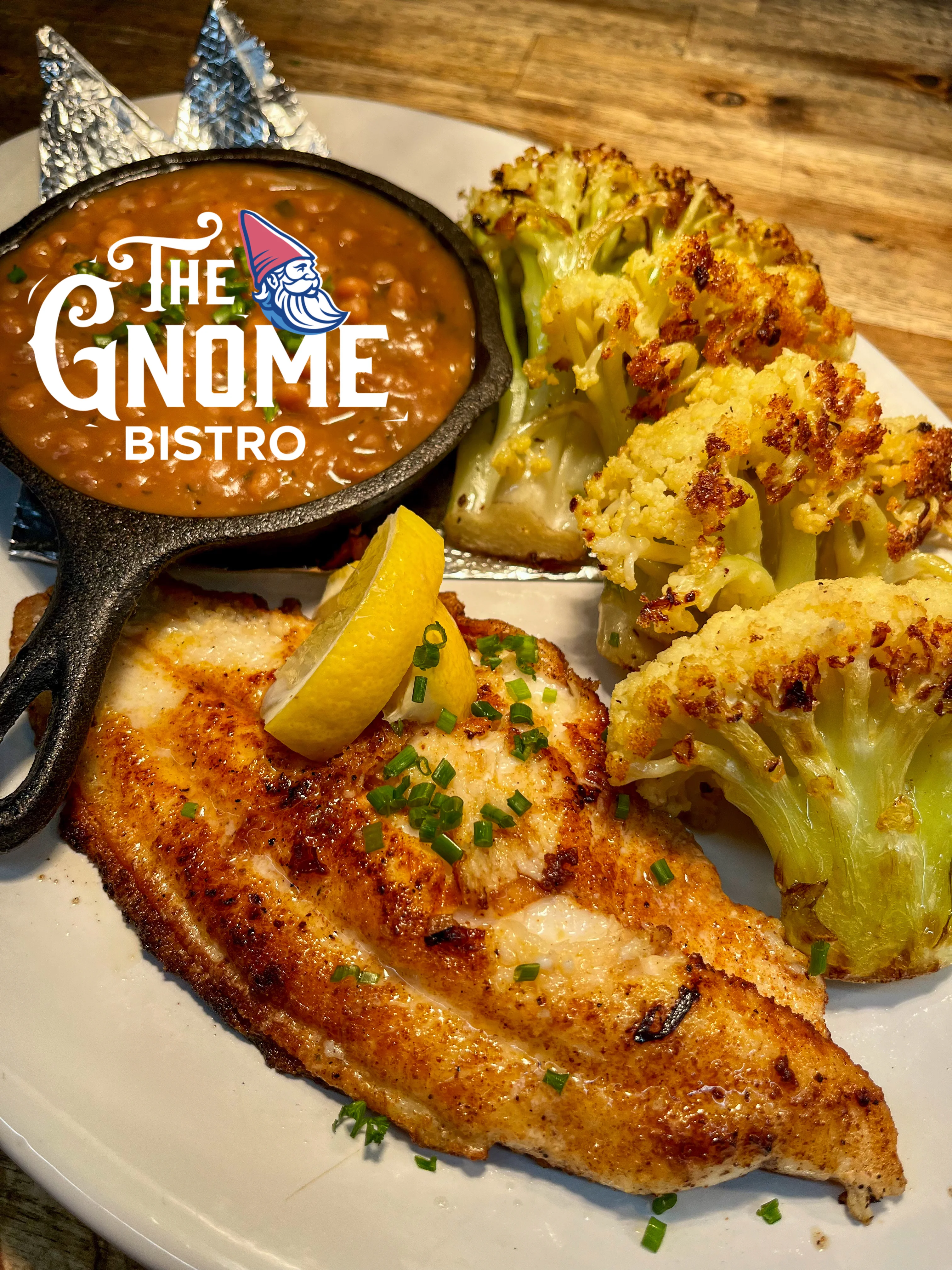 A beautifully plated dish featuring a fillet of fish alongside roasted cauliflower and a side of baked beans served in a small skillet. A lemon wedge and fresh chives add finishing touches.