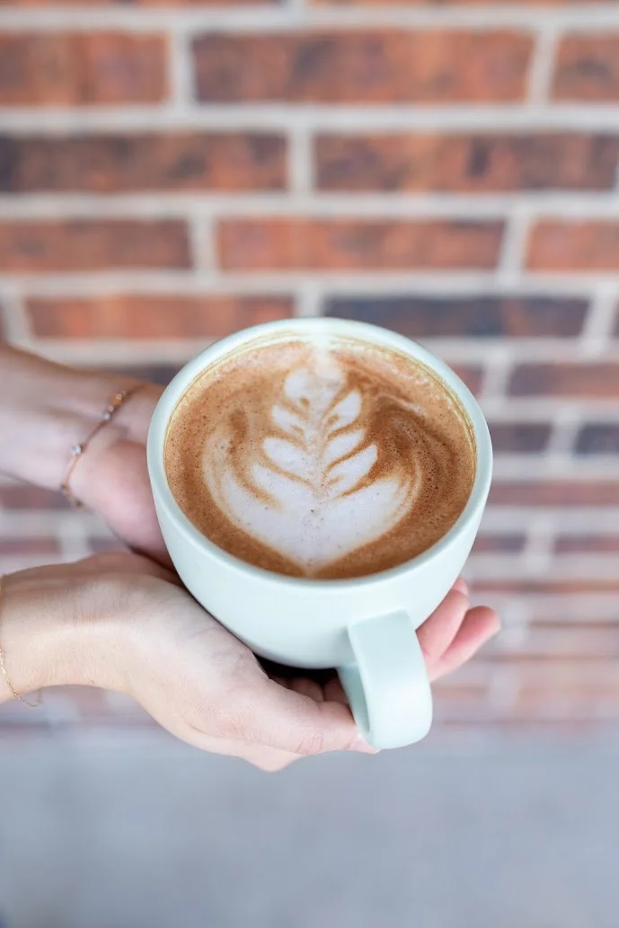 A steaming cup of coffee held with two hands, featuring a latte art design resembling a leaf on the surface. The background consists of a textured brick wall.
