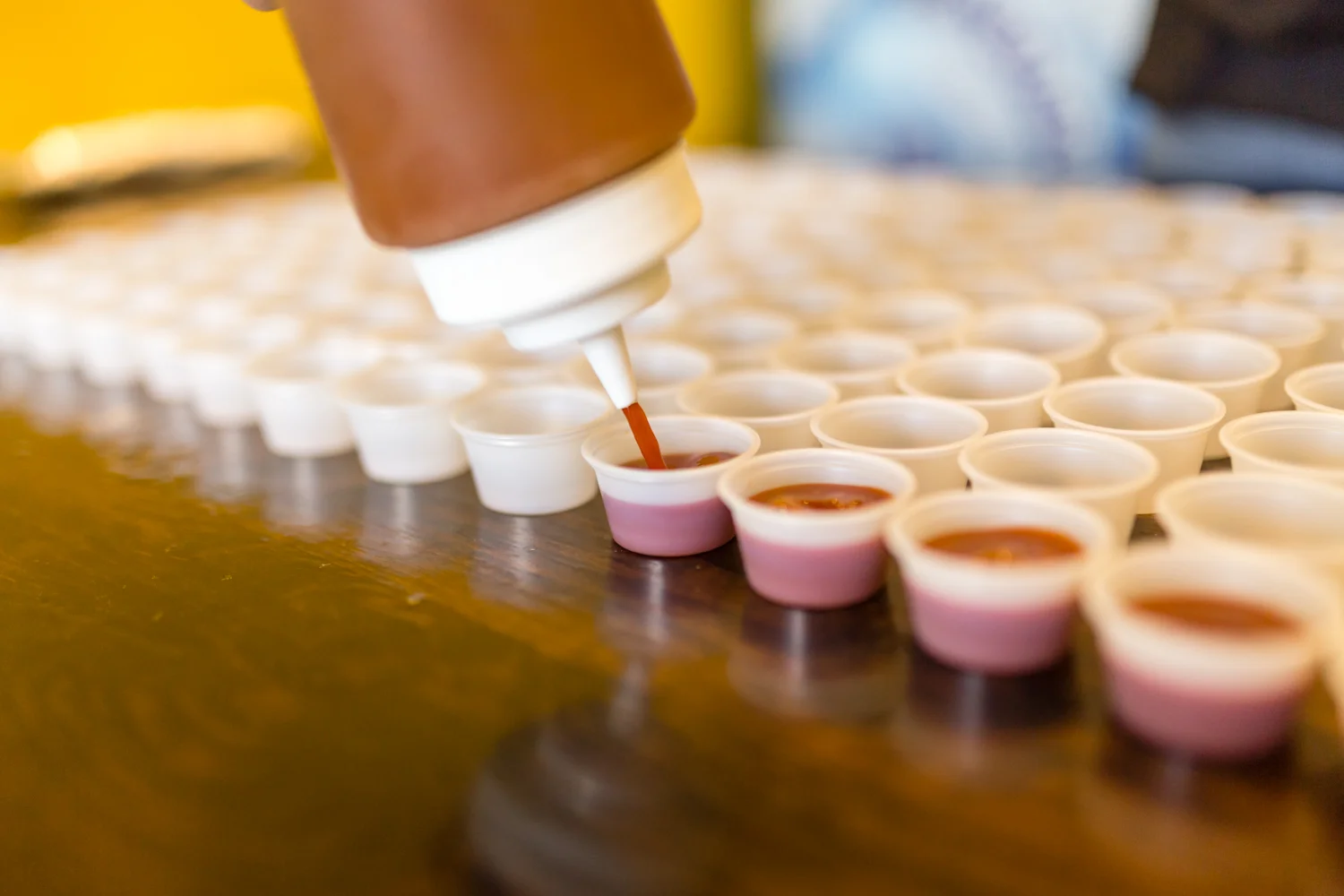 A squeeze bottle is dispensing sauce into small white cups arranged neatly on a wooden surface.