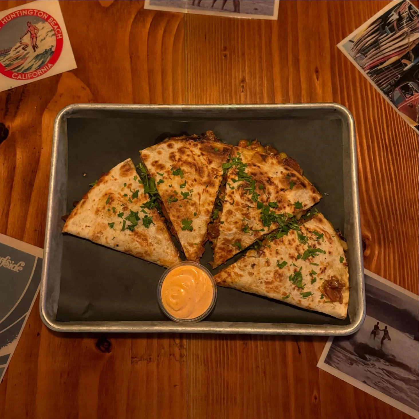 A plate of quesadillas cut into wedges, garnished with chopped parsley and served with a side of dipping sauce, placed on a wooden table.