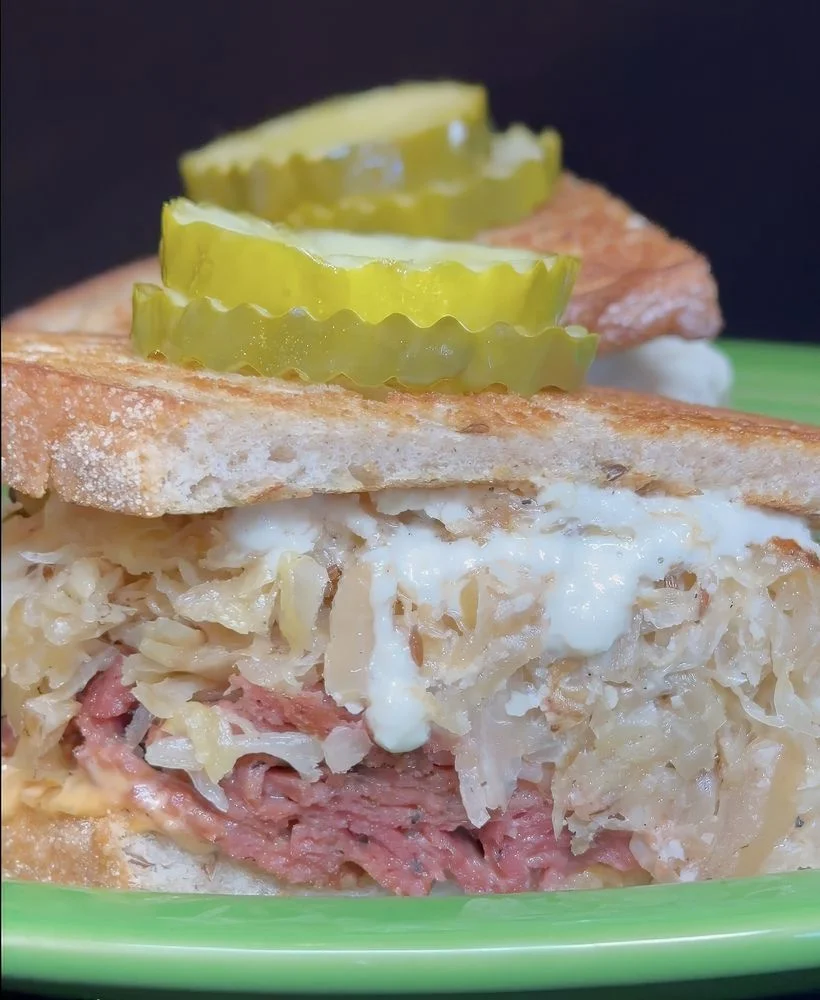 A sandwich featuring layers of meat, sauerkraut, and a creamy dressing, topped with sliced pickles on toasted bread.