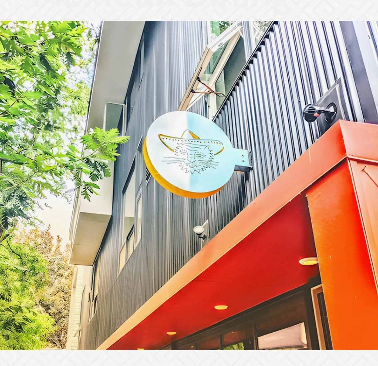 An outdoor view of a building with a distinctive sign featuring a stylized design, surrounded by greenery.