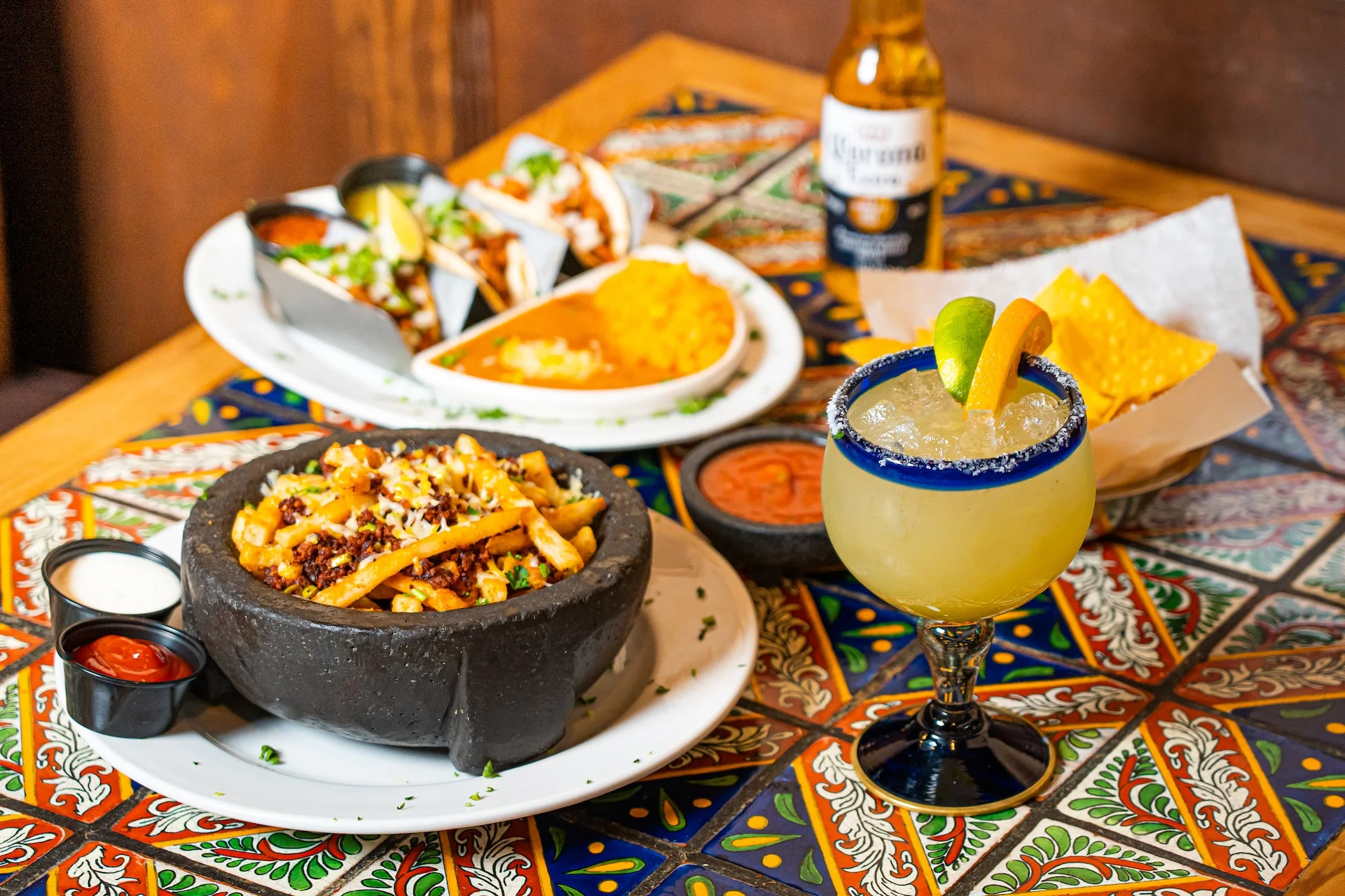 A colorful Mexican meal featuring a variety of dishes including loaded fries, tacos, rice, and drinks, all arranged on a vibrant table.