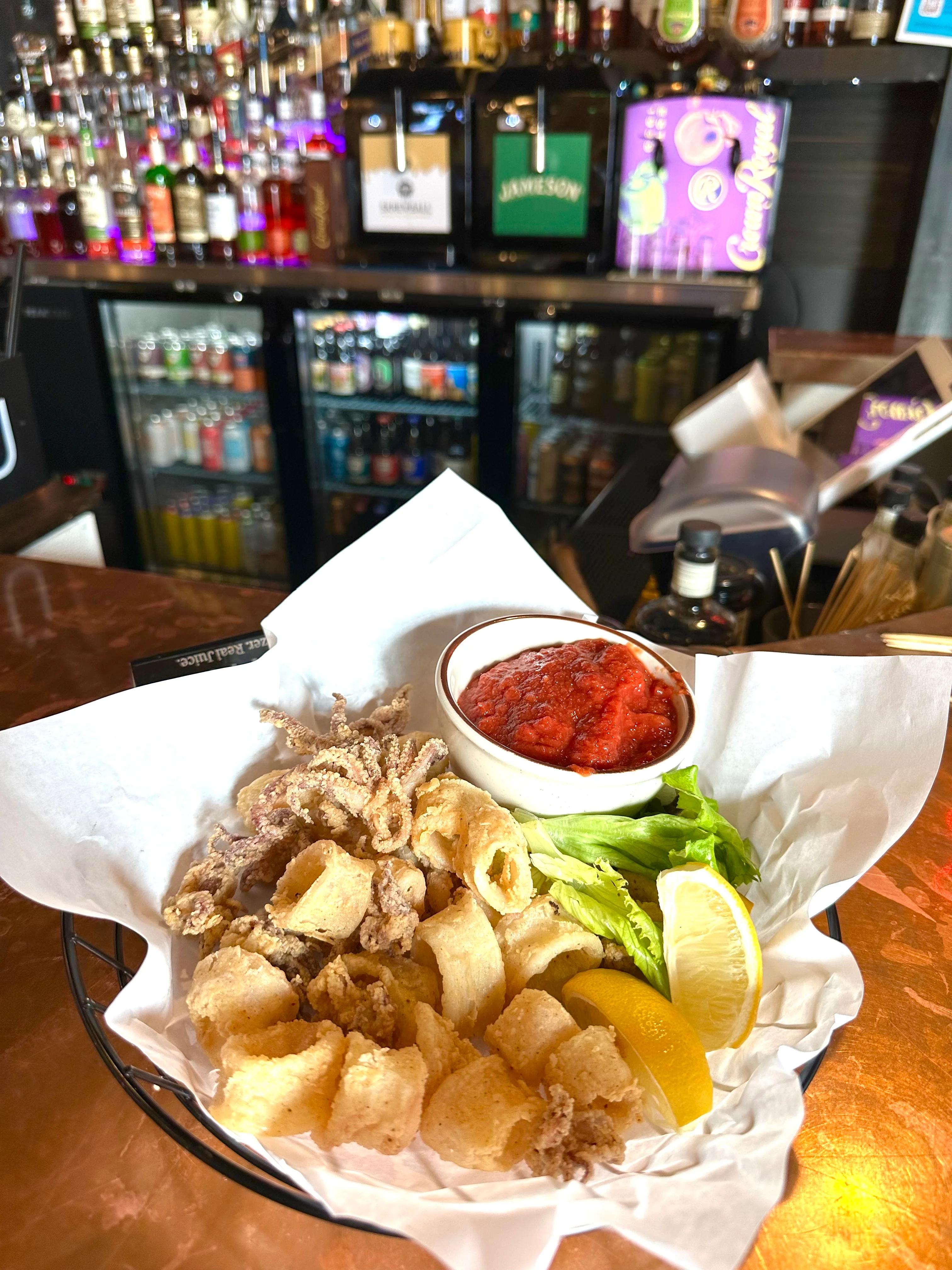 A serving of fried calamari with a side of marinara sauce, garnished with lettuce and lemon slices, arranged in a basket.