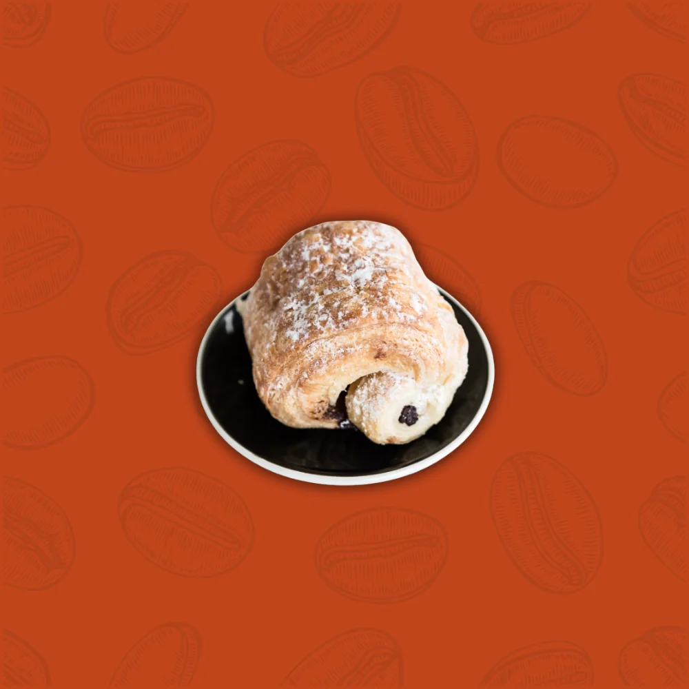 A flaky chocolate croissant dusted with powdered sugar sits on a black plate against an orange background with coffee bean patterns.