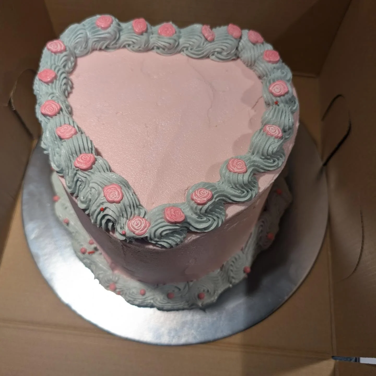 A heart-shaped cake adorned with pink and light green frosting flowers, resting inside a box.