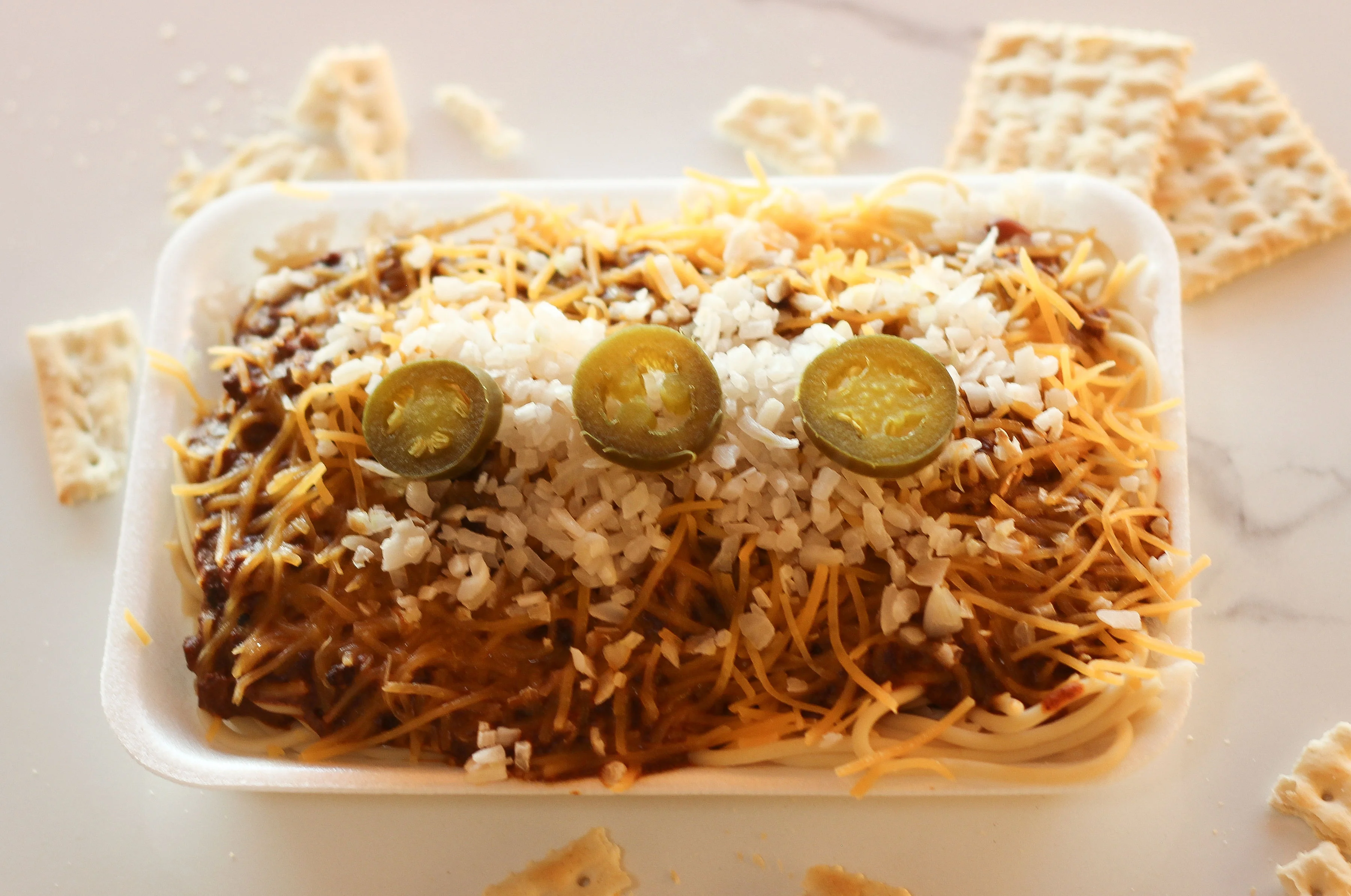A food container filled with spaghetti topped with chili, shredded cheese, diced onions, and sliced jalapeños, accompanied by snack crackers.