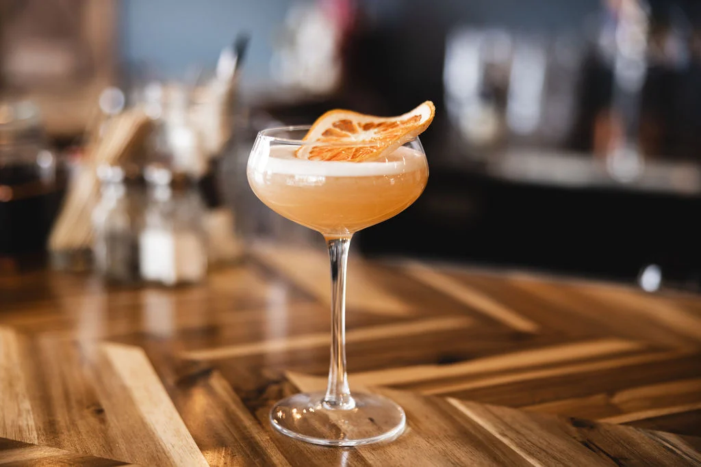 A handcrafted cocktail in a coupe glass garnished with a dried orange slice, set on a wooden table.