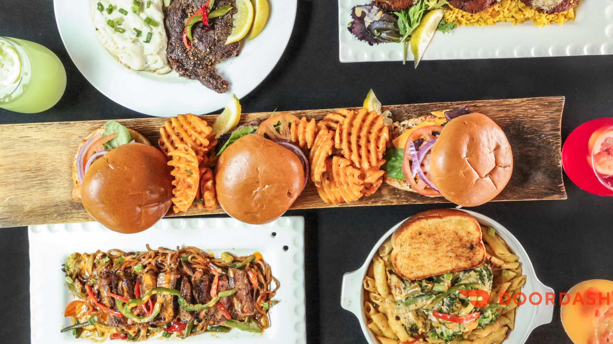 A variety of dishes featuring burgers, pasta, and sides on a black table. The presentation includes two burgers with waffle fries, a stir-fry dish, and a pasta bake with toast.