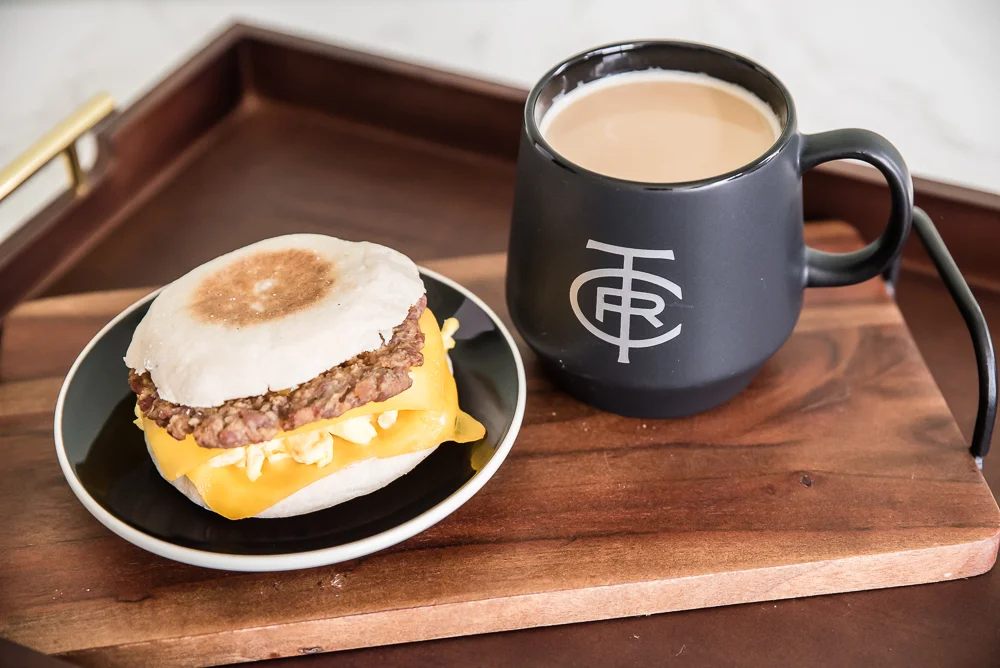 A breakfast sandwich with cheese on a small plate, next to a black mug filled with coffee on a wooden tray. Warm, cozy tone.