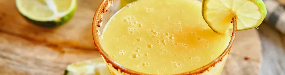 Two glasses of yellow drink with lime slices and chili rim on a wooden surface.