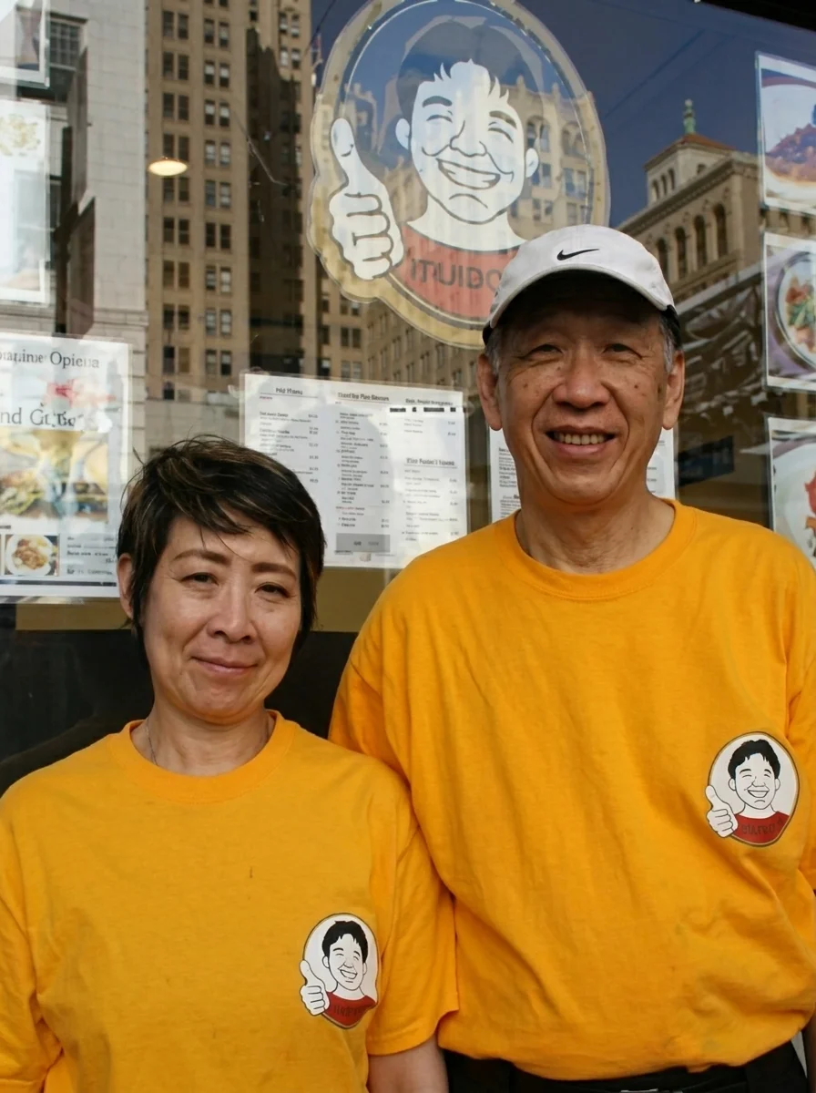 Muracci's founders, husband-and-wife team Yasuyuki and Tamiko Murata, wearing matching yellow Muracci's T-shirts and smiling in front of the restaurant's window display.