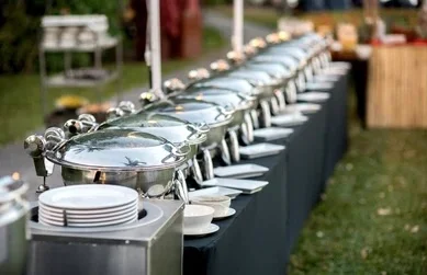 A long buffet table set outdoors with multiple silver warming trays and neatly arranged plates ready for serving.