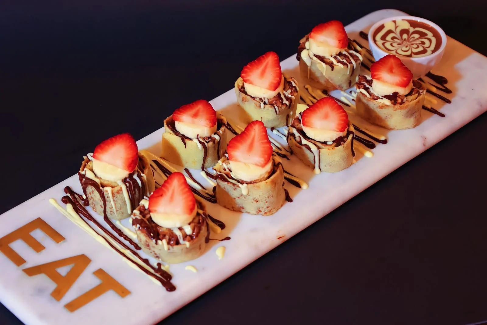 A colorful serving of rolled desserts topped with strawberries and bananas, drizzled with chocolate and white sauces on a decorative plate.