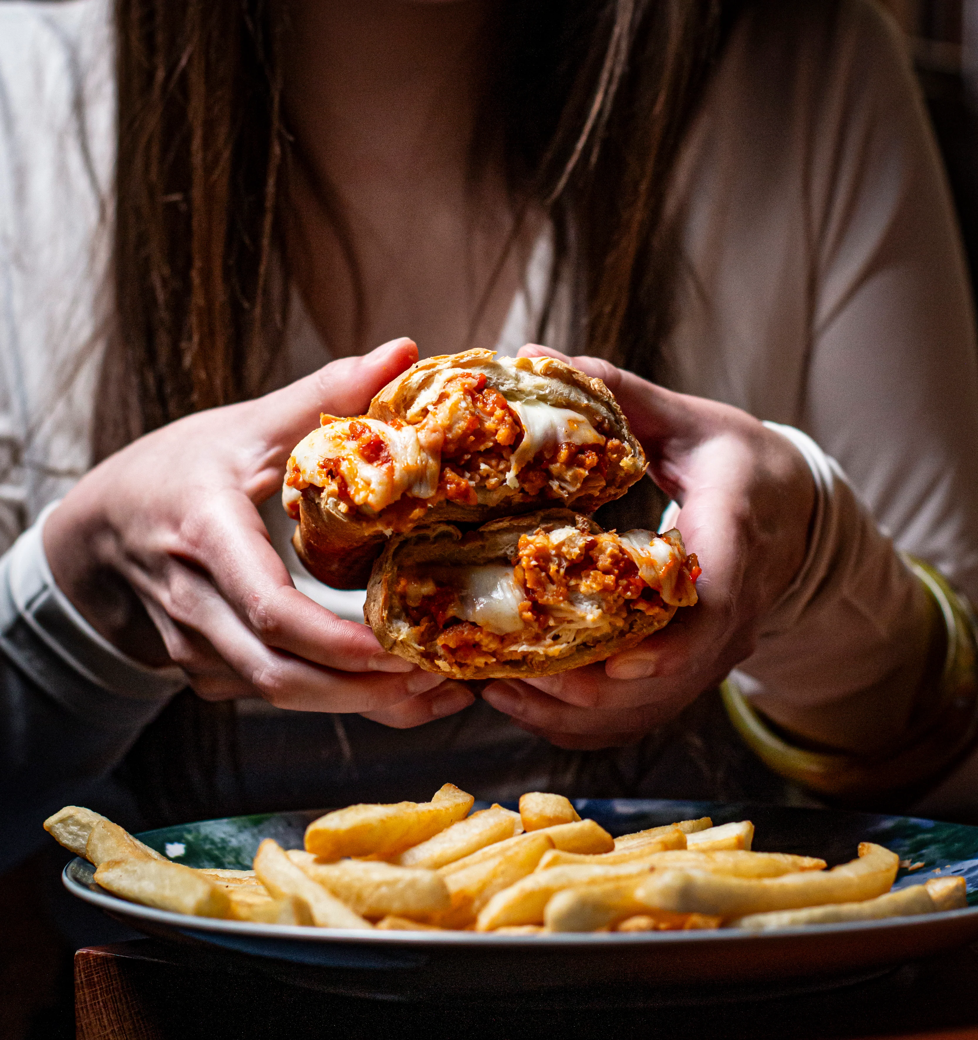 A person holding two halves of a sandwich filled with chicken and cheese, accompanied by a side of fries.