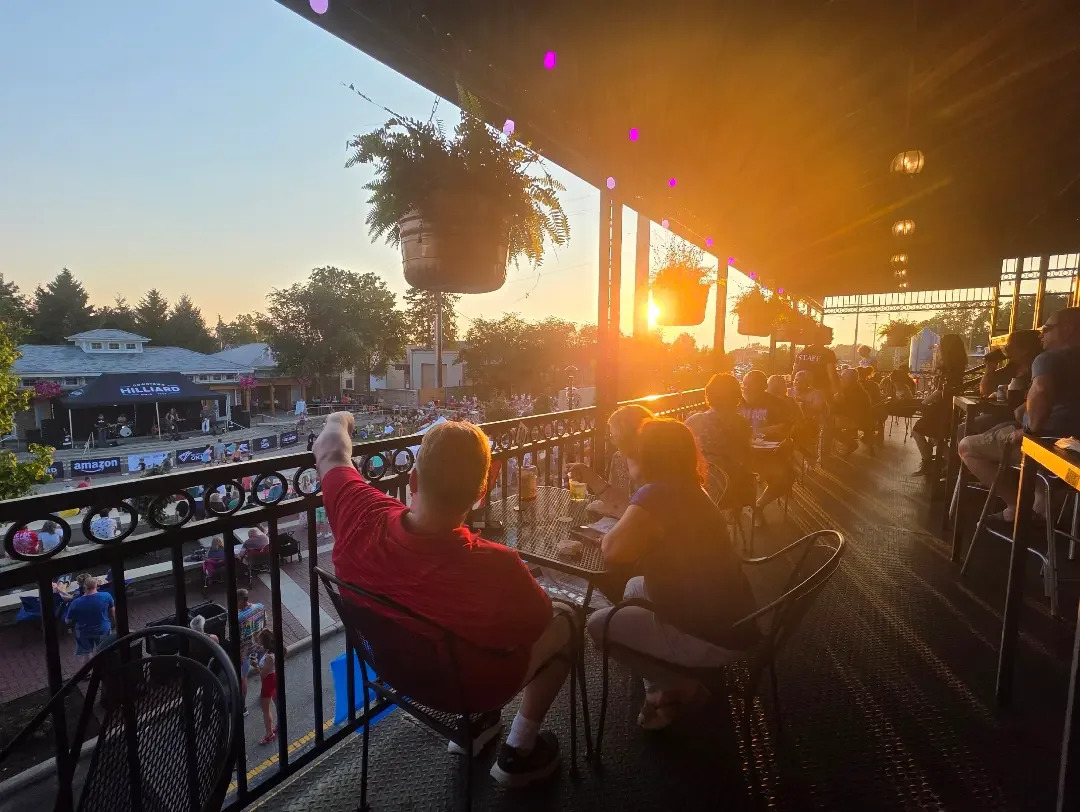 An outdoor seating area with guests enjoying the sunset, overlooking a lively street event. The warm light creates a festive atmosphere.