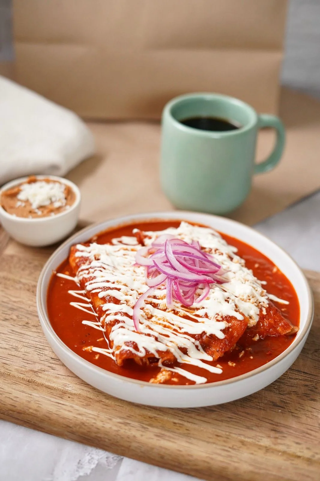 A plate of enchiladas topped with crema, cheese, and red onion slices, served with a side of refried beans and a cup of coffee.