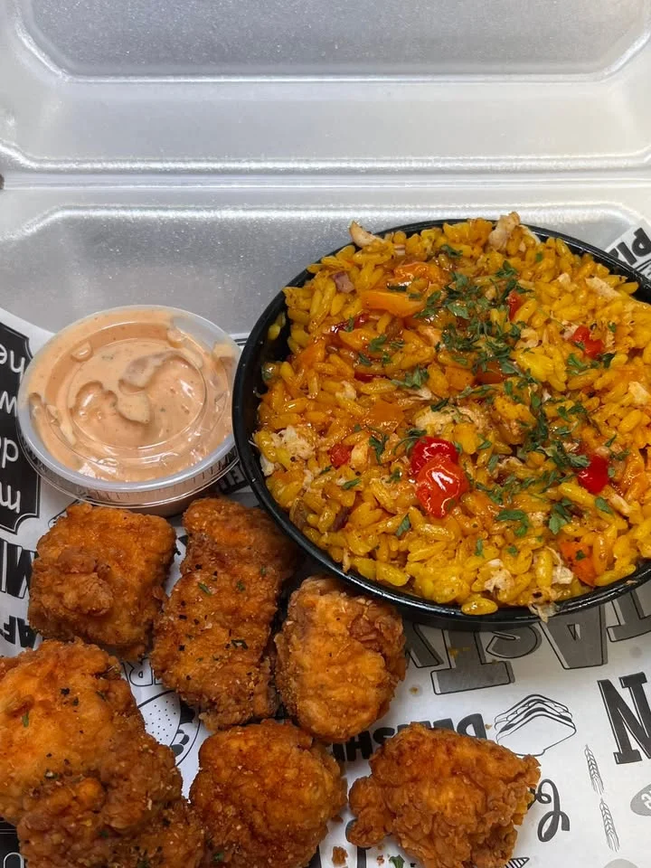 A serving of seasoned rice accompanied by fried chicken pieces and a dipping sauce. The food is presented in a takeout container.