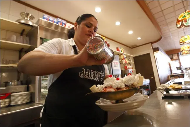 A server decorates a bowl of ice cream with whipped cream and colorful sprinkles in a diner setting.
