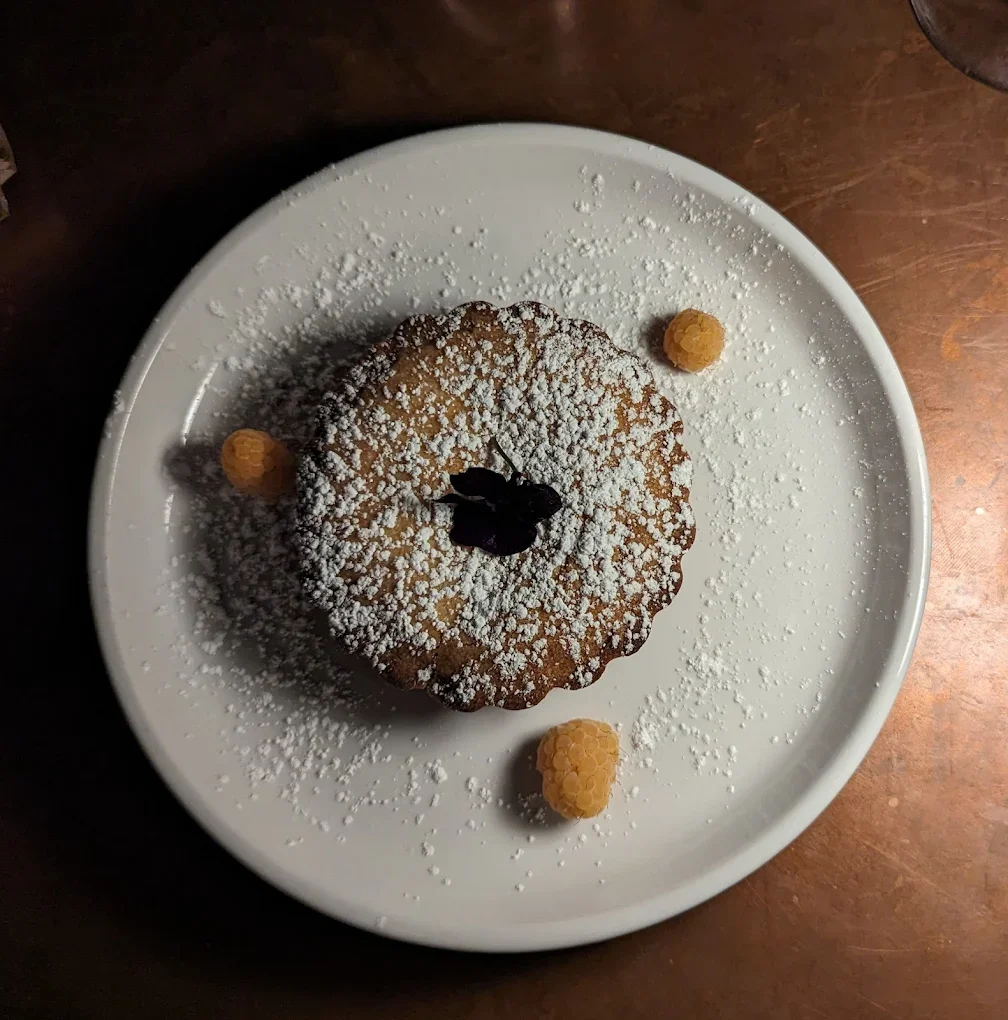 A round dessert dusted with powdered sugar, featuring a flower garnish, surrounded by small fruit garnishes on a white plate.