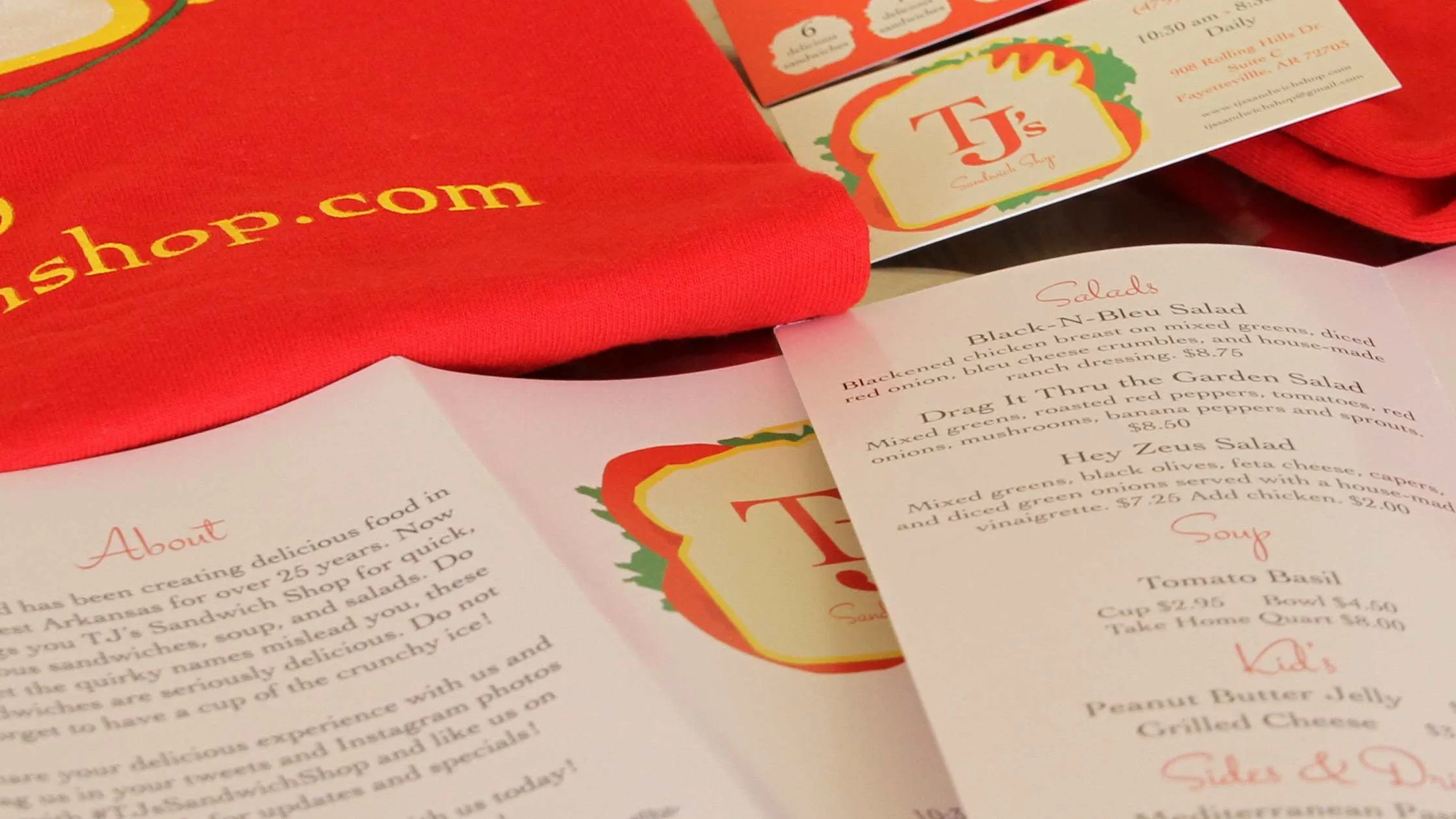 Close-up of TJ's Sandwich Shop promotional materials, including printed menus and flyers with colorful sandwich-themed branding. A red T-shirt featuring the shop's website is partially visible. The menu highlights salads like 'Black-N-Bleu' and 'Hey Zeus Salad,' along with soups and kids’ items.