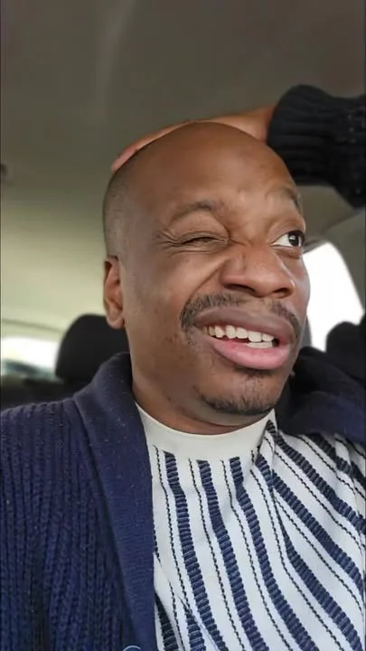 A person making a facial expression while sitting in a car, with hands on their head and a playful look.