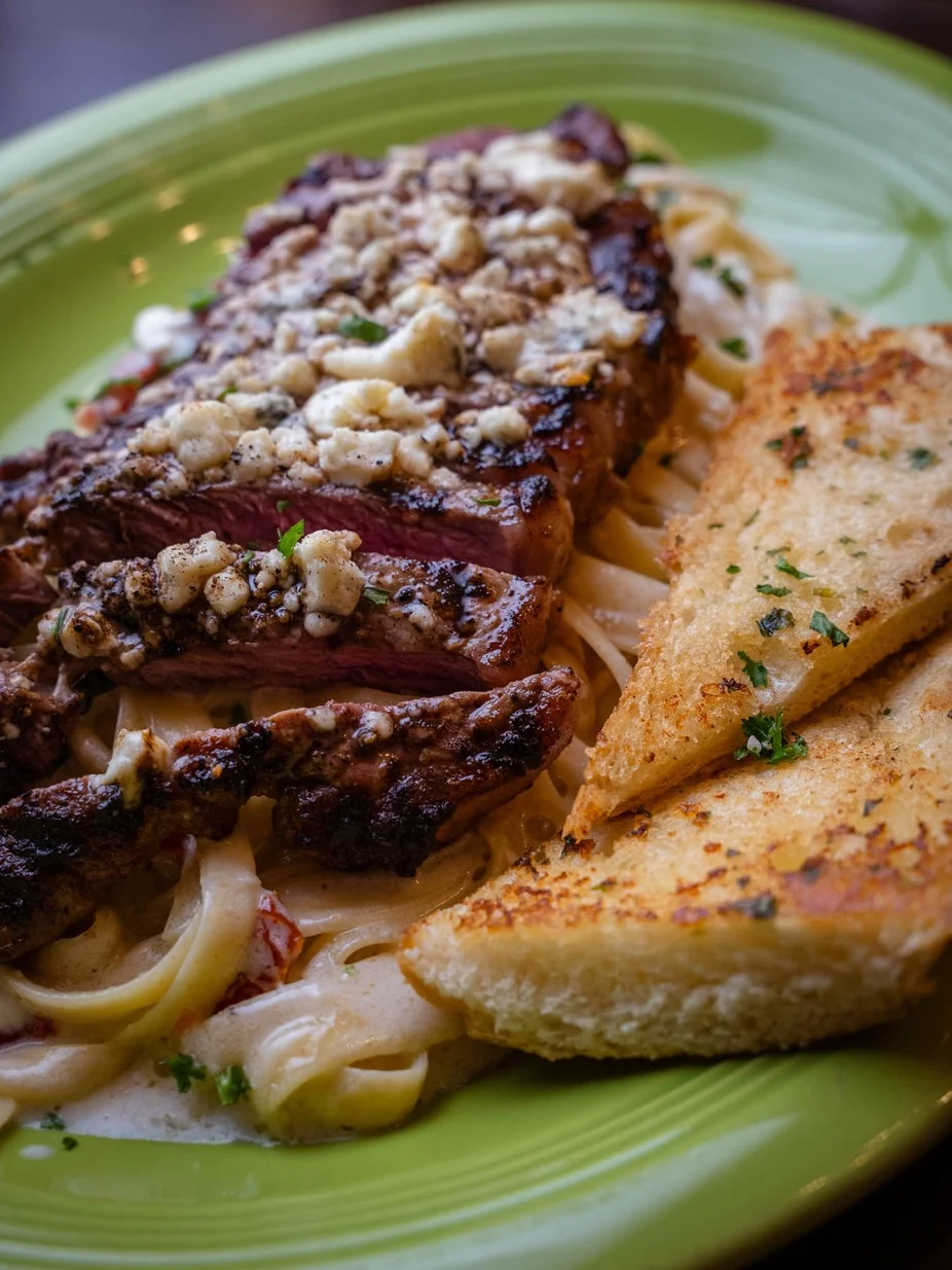 A hearty meal featuring grilled steak topped with crumbled cheese, served on a bed of fettuccine pasta in a creamy sauce, accompanied by toasted garlic bread.