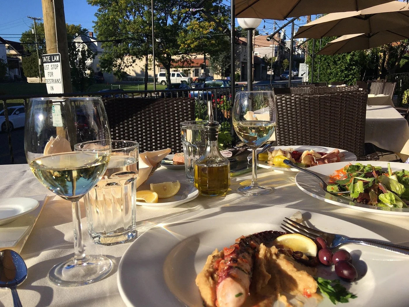 Outdoor patio dining at Toscana on Main in Westchester. Dining scene with elegantly set table, featuring plates of seafood and salad. Glasses of white wine and water, sunlight casting soft shadows.