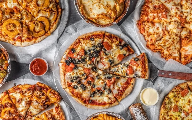 A spread of various pizzas, including a sliced pizza topped with tomatoes, olives, and cheese, alongside side dishes and a dipping sauce.
