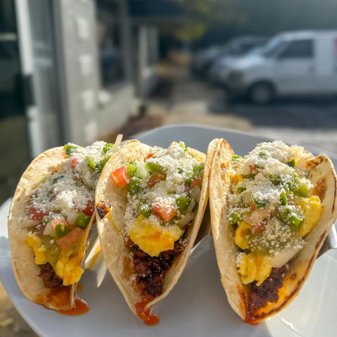 Three soft tacos filled with scrambled eggs, ground meat, and topped with a fresh mix of diced peppers, tomatoes, and cheese.