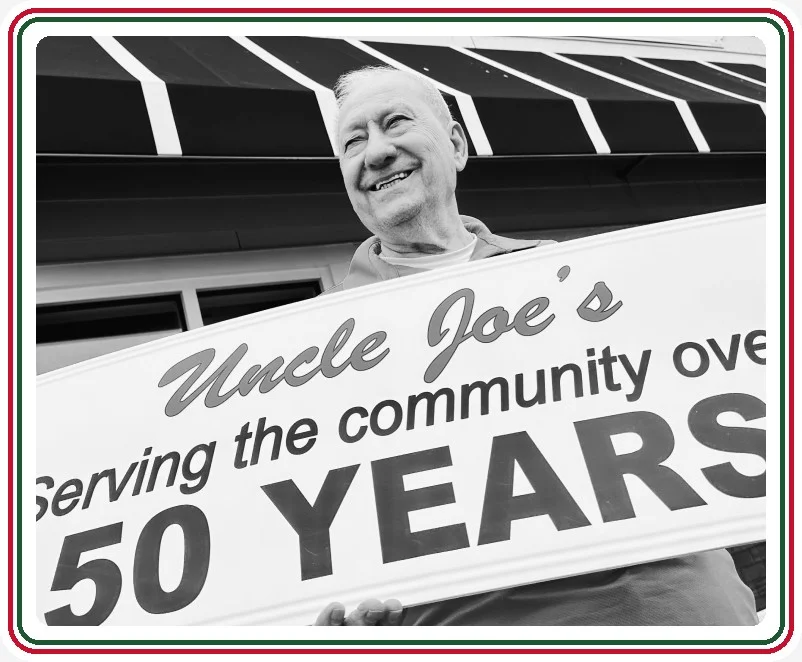 Uncle Joe outside the shop on Montauk Highway, holding a sign that reads "Uncle Joe's: Serving the community over 50 years"