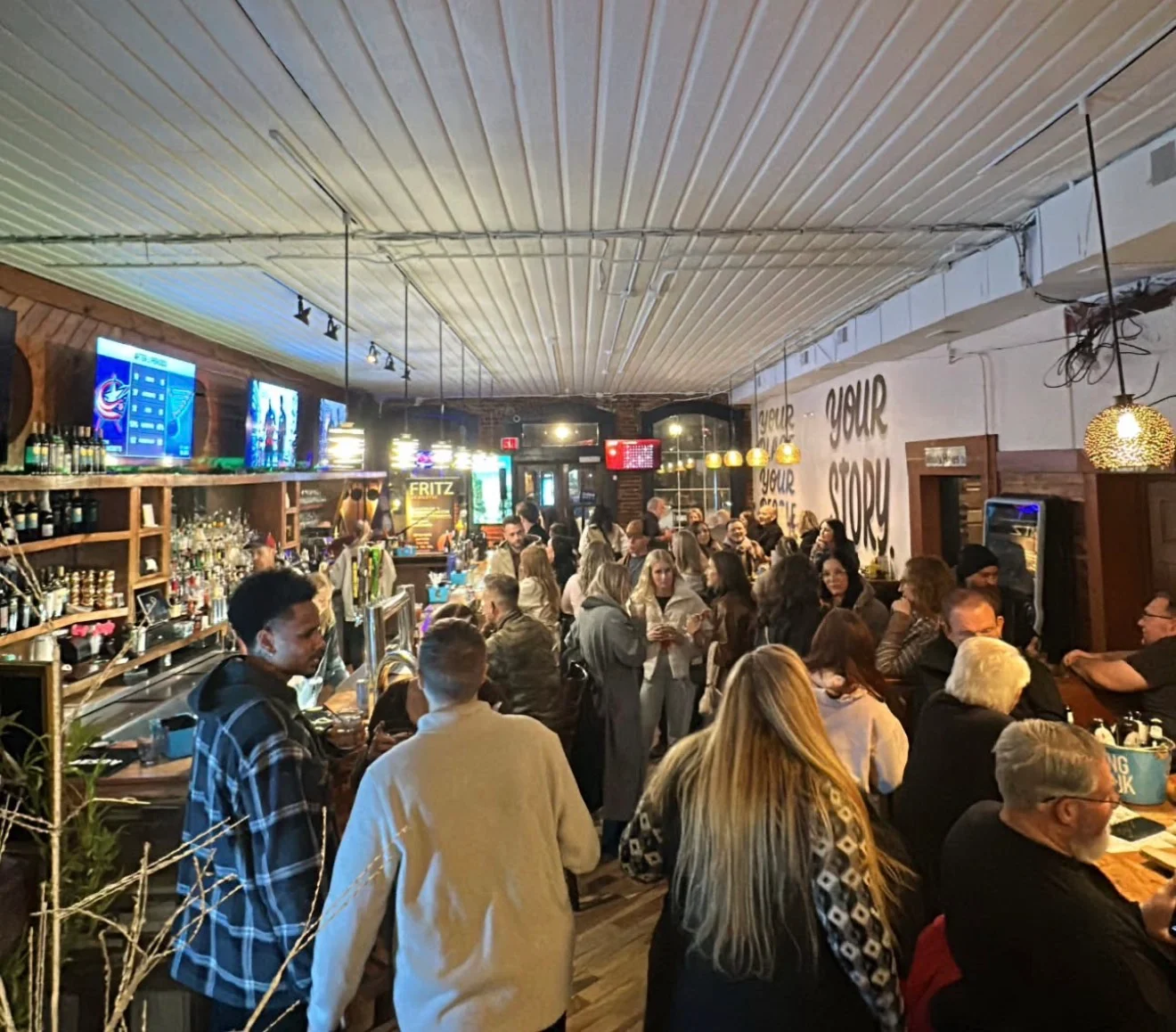 A lively bar scene filled with patrons, featuring a long bar stocked with drinks, multiple screens displaying information, and a warm, inviting atmosphere.
