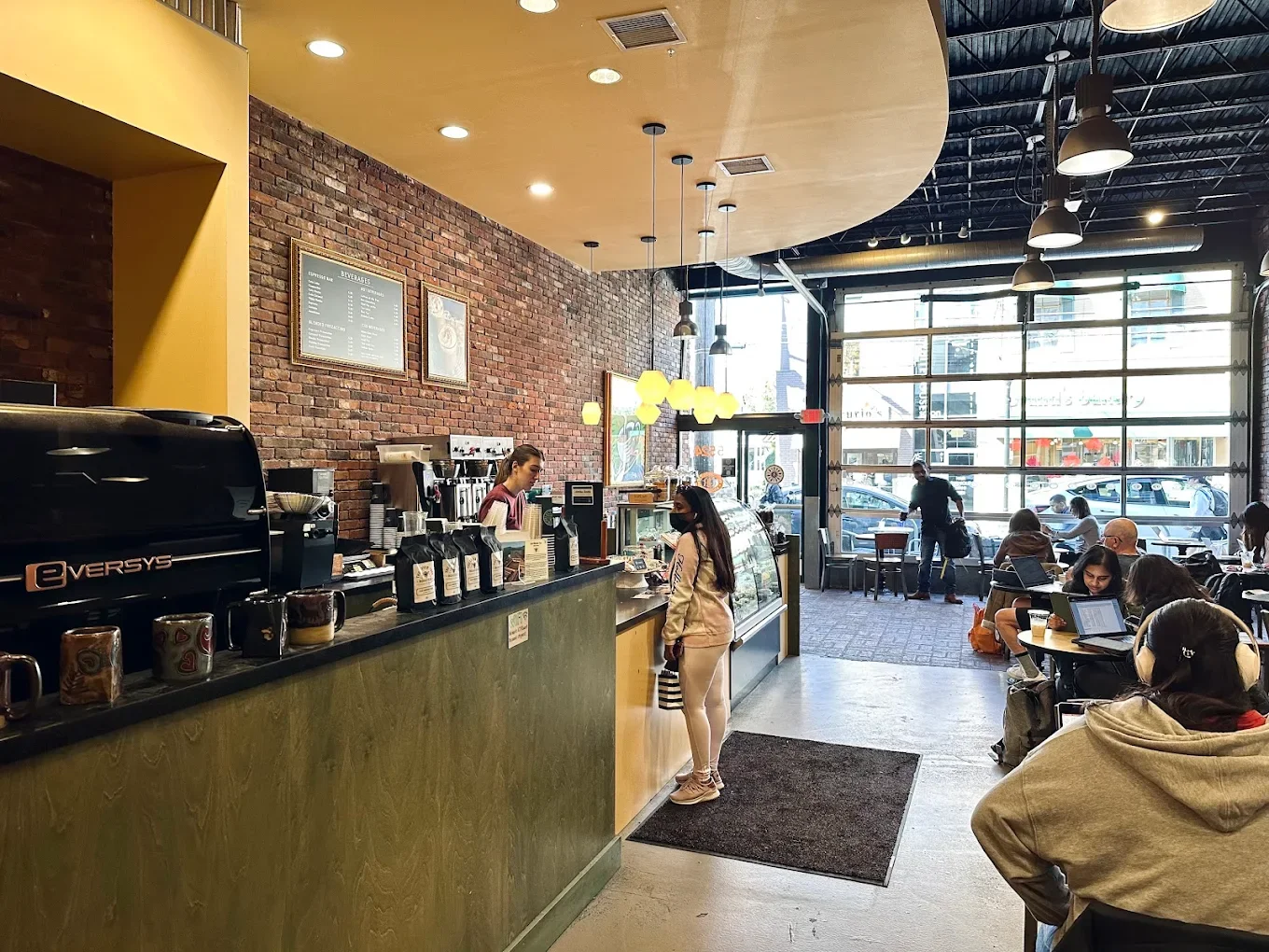 A cozy café with warm lighting and a brick wall. A customer orders at the counter, while others sit at tables using laptops, creating a busy, relaxed atmosphere.