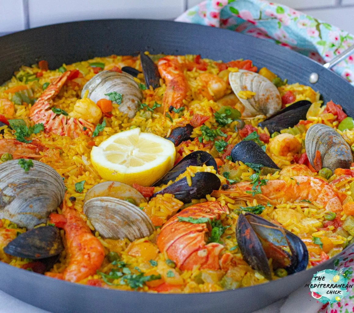 A large paella dish filled with rice, shrimp, clams, mussels, and colorful vegetables, topped with a slice of lemon and herbs.