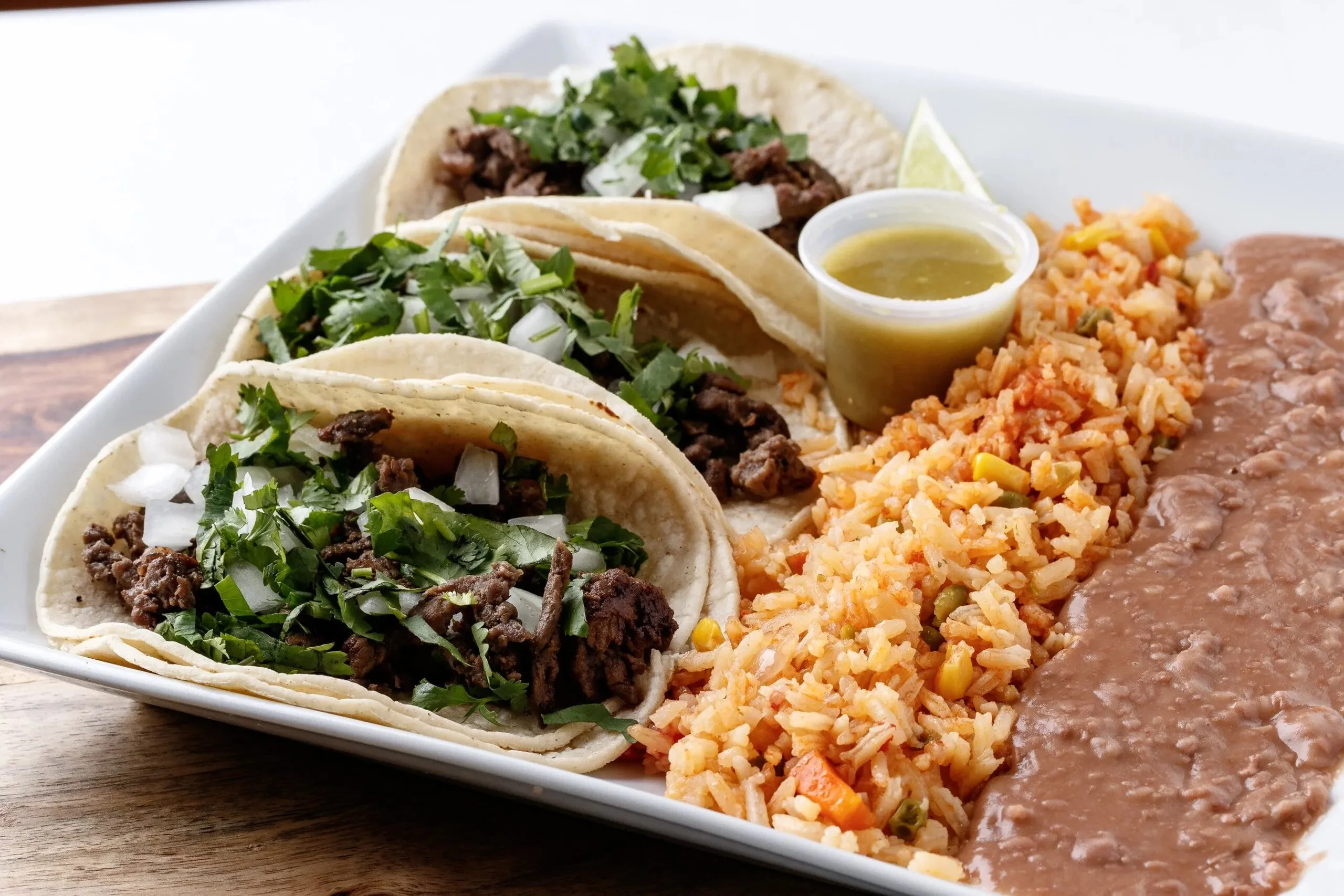 A plate of three tacos filled with meat, topped with chopped cilantro and onions, served alongside rice, beans, and a small container of sauce.