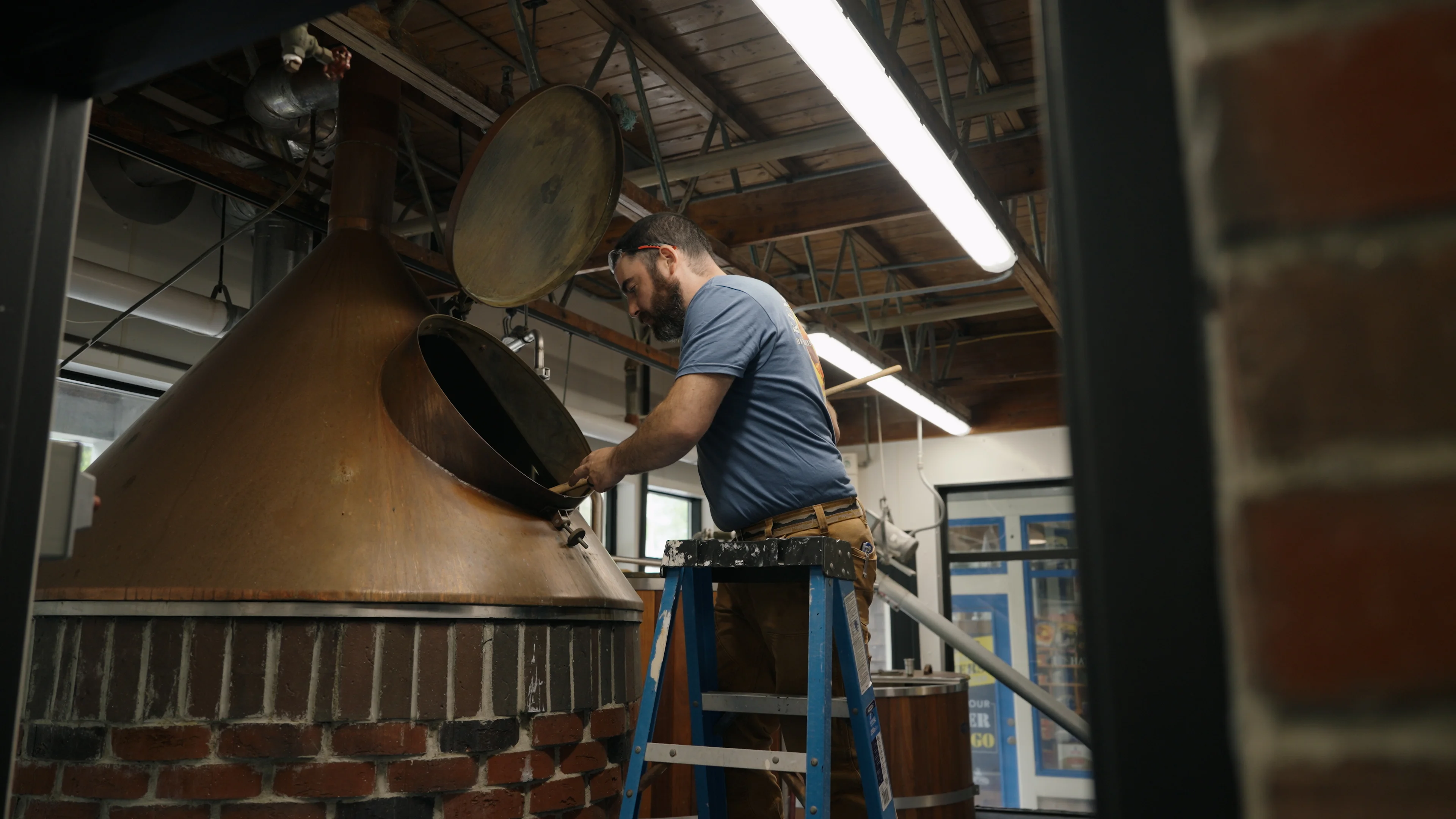 A person working on brewing equipment inside a brewery. A metal kettle is being inspected, and the setting features industrial-style decor.