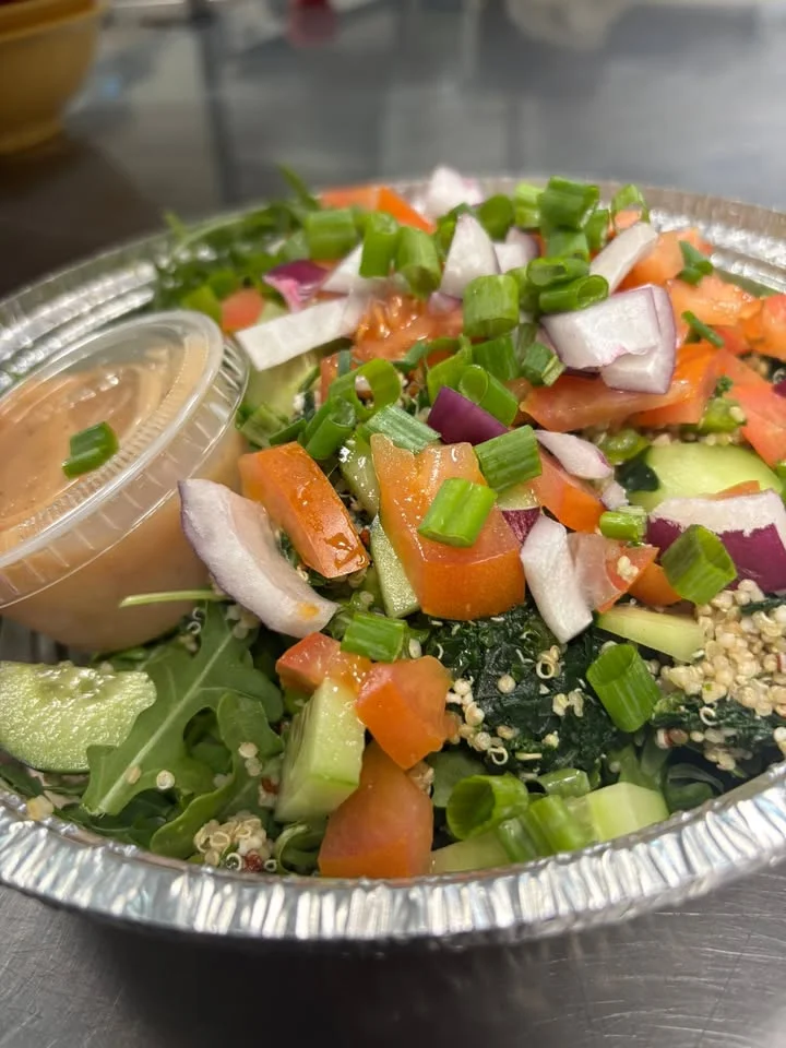 A vibrant salad in a foil container, topped with fresh vegetables including tomatoes, cucumbers, and onions, served with a side of dressing.