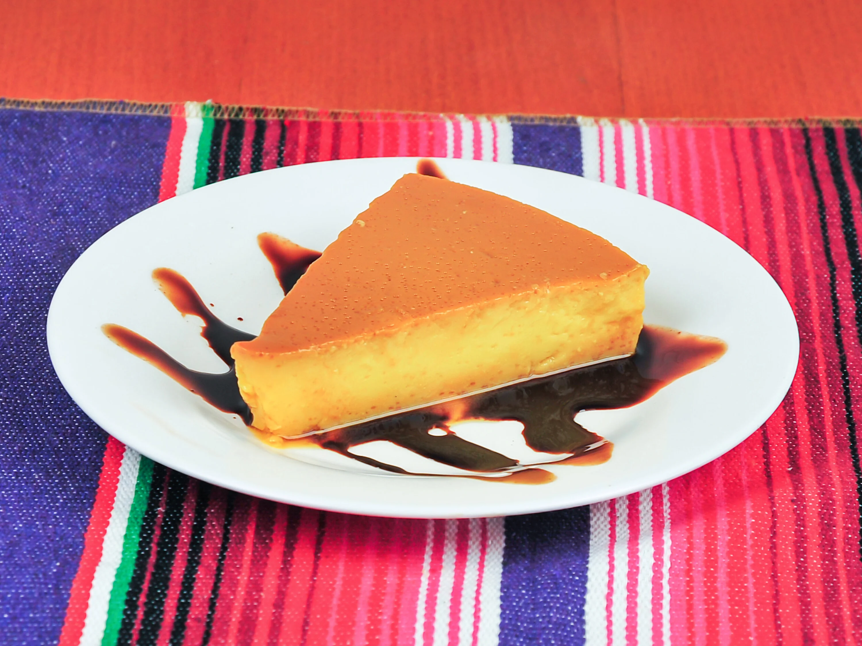 A slice of flan drizzled with a dark sauce, presented on a white plate. The dish is placed against a bright striped background.