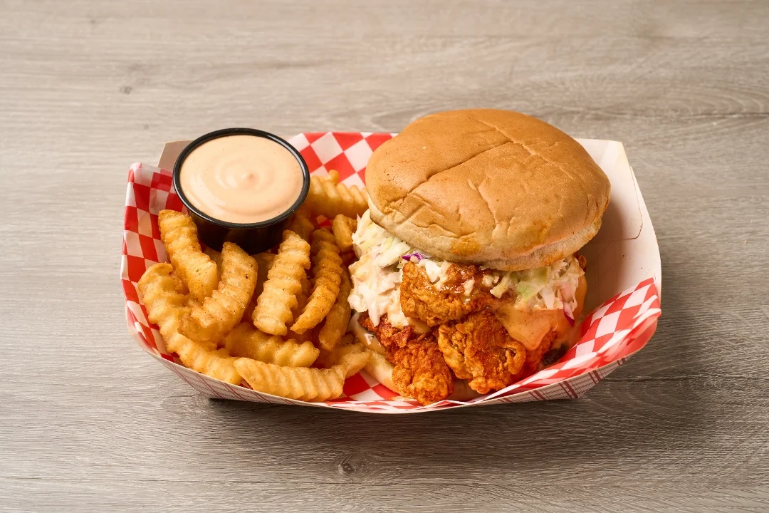 A chicken sandwich served with coleslaw in a bun, accompanied by crispy crinkle-cut fries and a small container of dipping sauce.