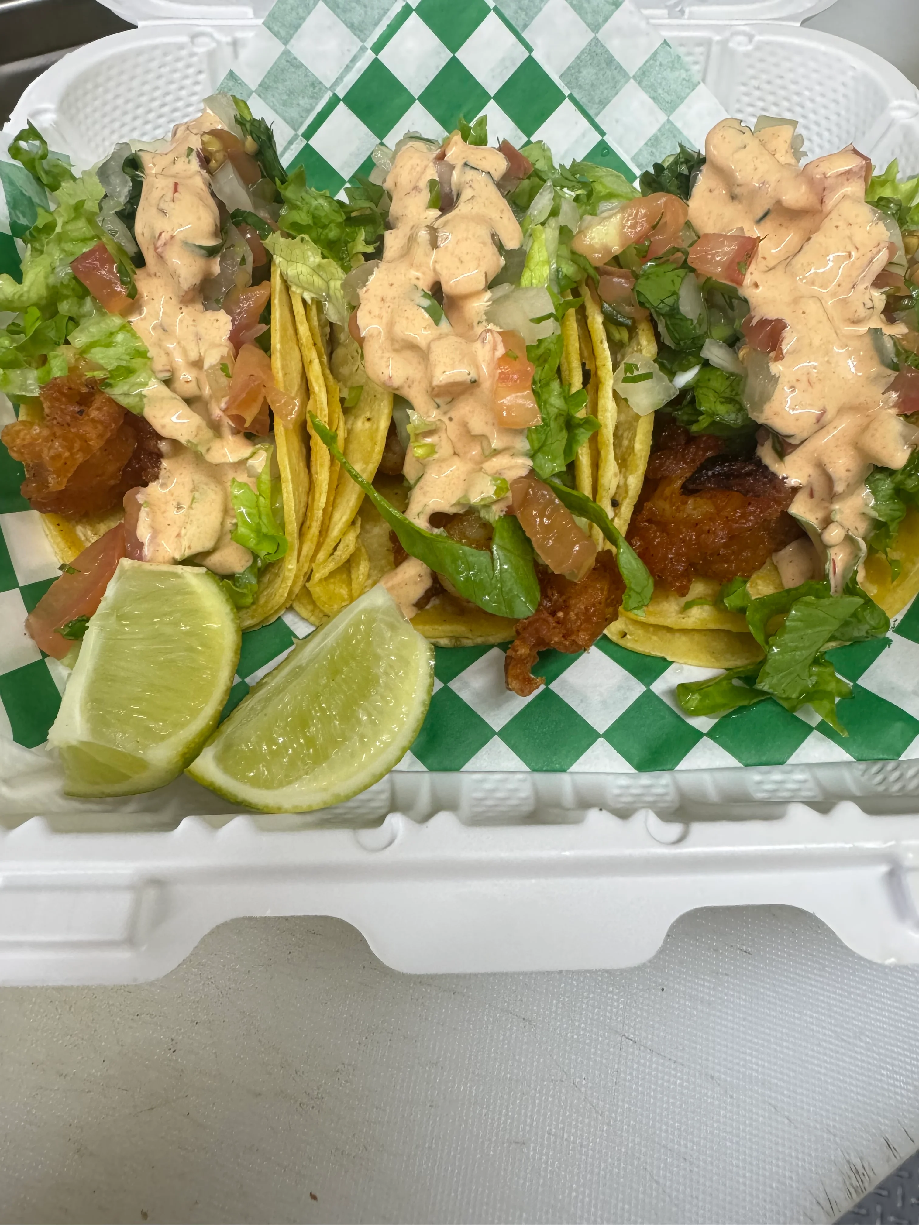 A serving of three tacos topped with lettuce, tomatoes, and a creamy sauce, along with lime wedges, presented in a takeout container.