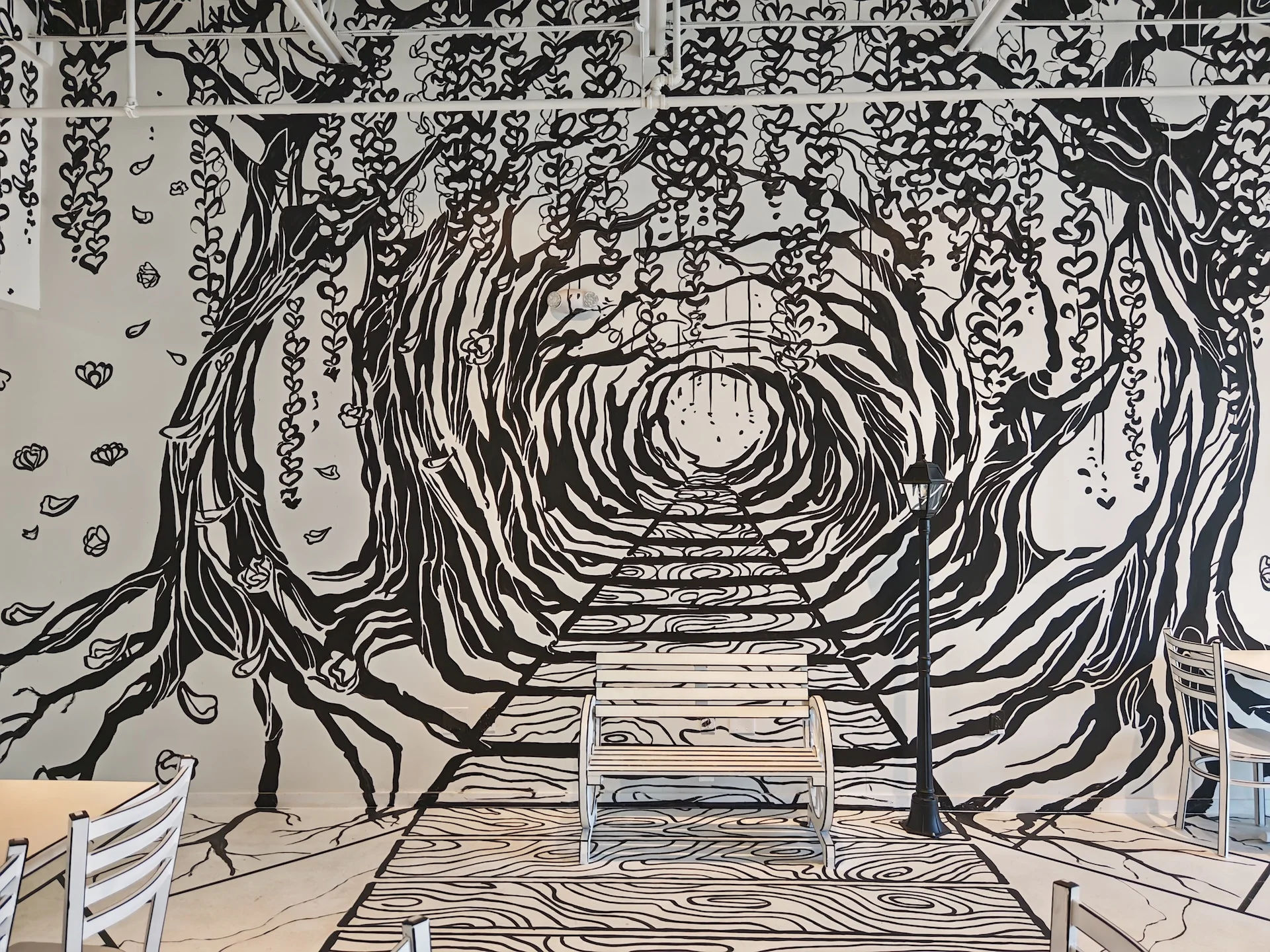 An immersive, black and white wall mural at Chopsticks Charm that creates an illusion of a path leading into a tunnel of trees, complete with a 2-D style bench.