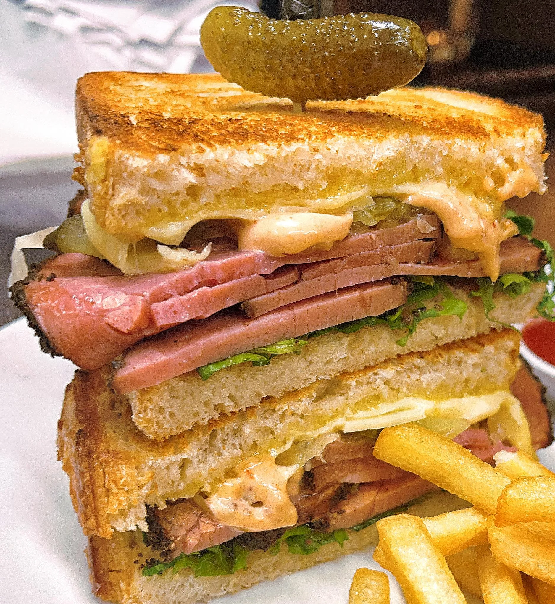A towering sandwich stacked with layers of sliced meat, lettuce, and sauce, topped with a pickle on toast. Served alongside crispy fries.