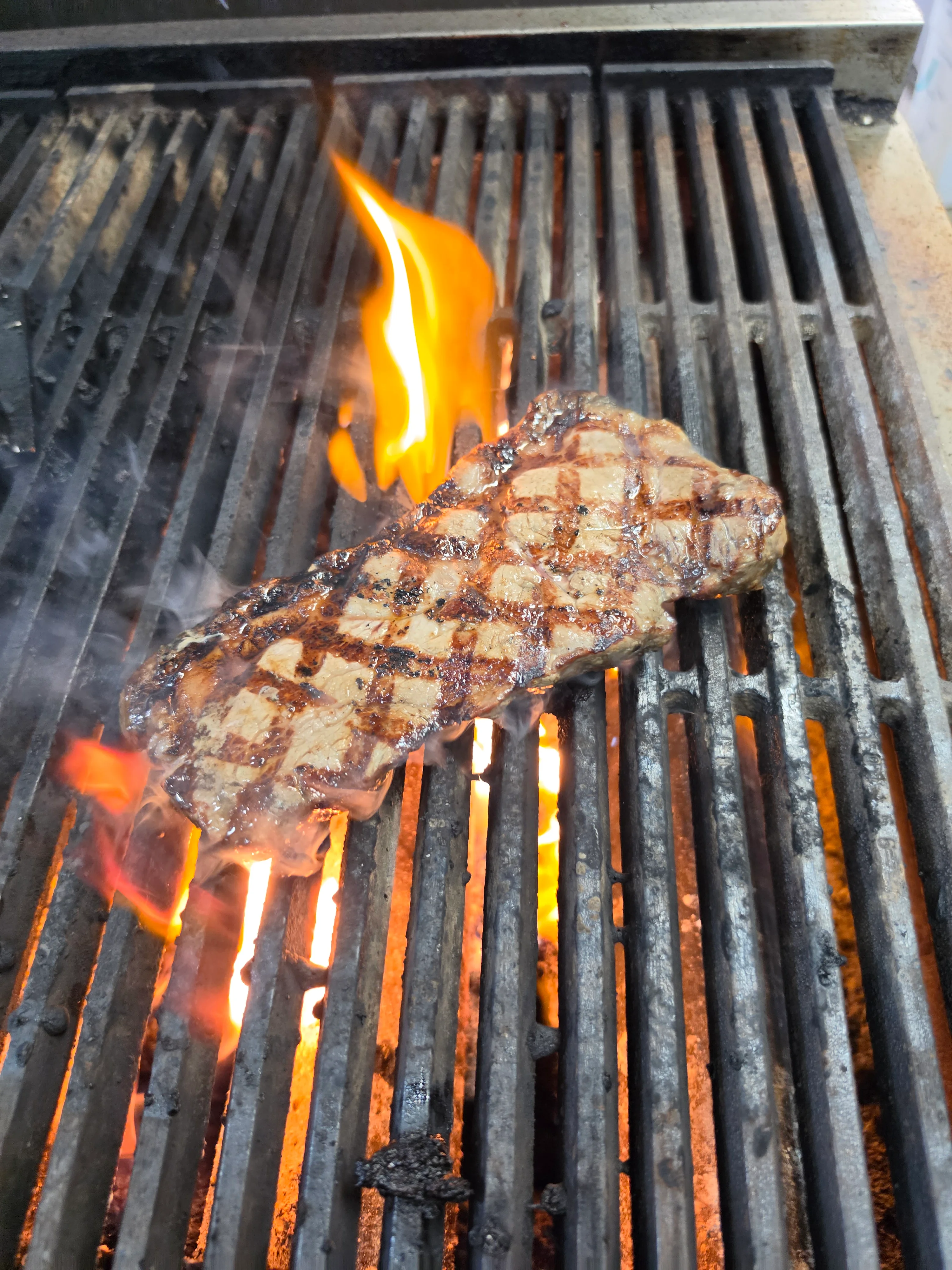 A steak sizzling on a grill with flames and smoke rising. The grill marks are clearly visible, showcasing the cooking process.