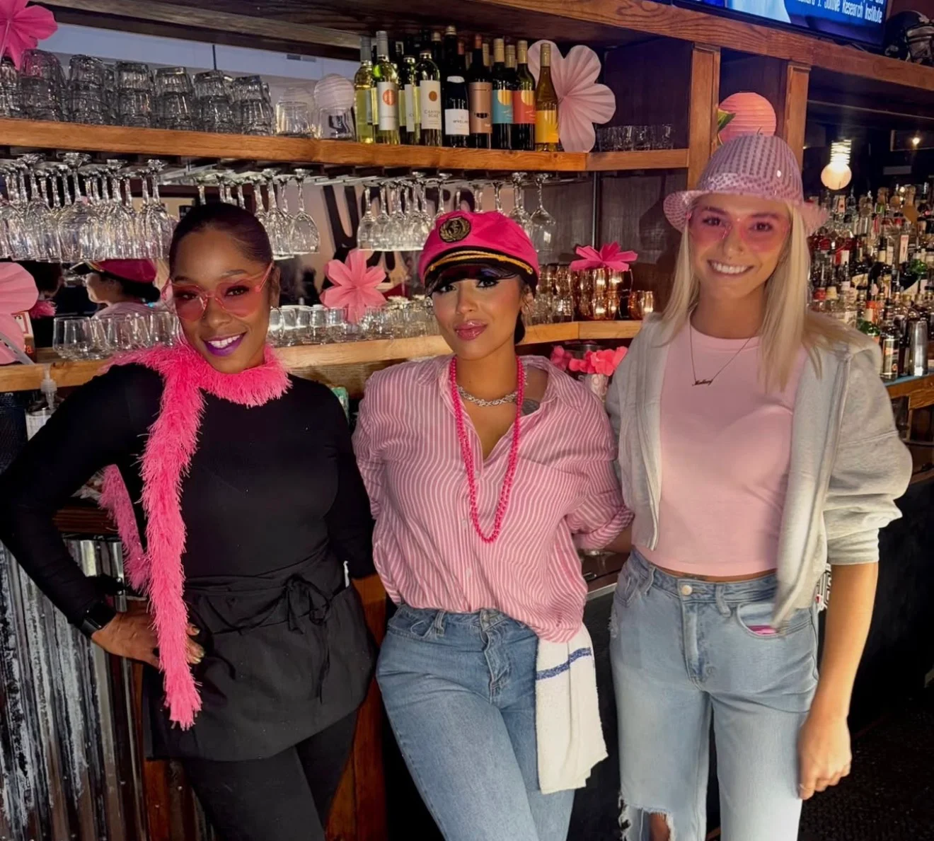Three individuals dressed in vibrant pink attire pose in front of a bar filled with various drinks and decorations.